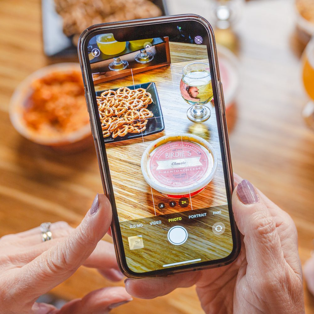 Hands photographing a tub of Birdie's Pimento Cheese and pretzels with a phone